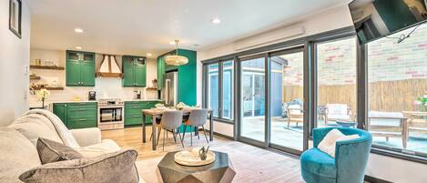 A modern living room with a sofa, a coffee table, and a blue armchair. There is a dining area with a table and chairs, and a kitchen with green cabinets.