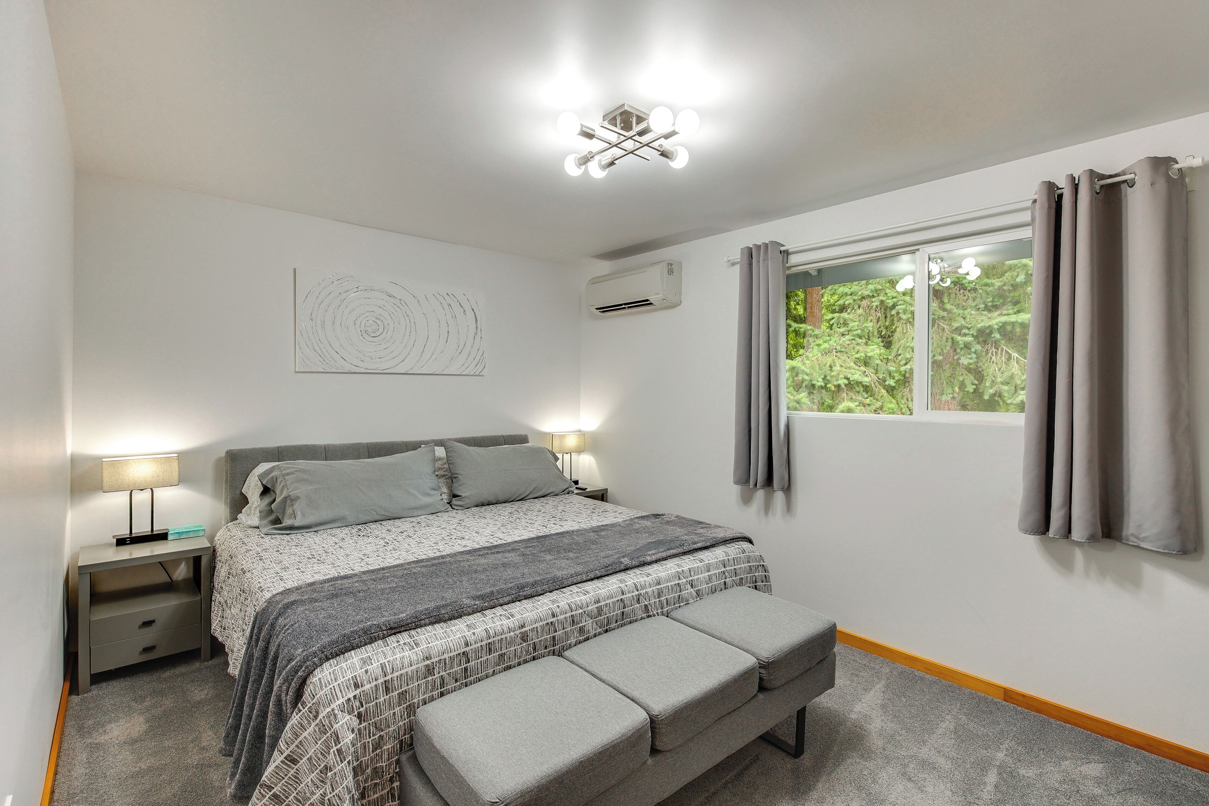 A bedroom with a bed, bedside lamps, a window with curtains, an air conditioning unit, and a wall-mounted artwork.
