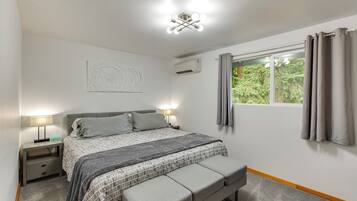 A bedroom with a bed, bedside lamps, a window with curtains, an air conditioning unit, and a wall-mounted artwork.
