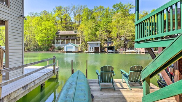 Une jetée en bois avec un canoë, deux fauteuils Adirondack verts et une vue sur un lac, avec des bâtiments en arrière-plan.