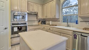 Una cocina con armarios de madera clara, electrodomésticos de acero inoxidable, una isla grande y un ventanal con vista a árboles.