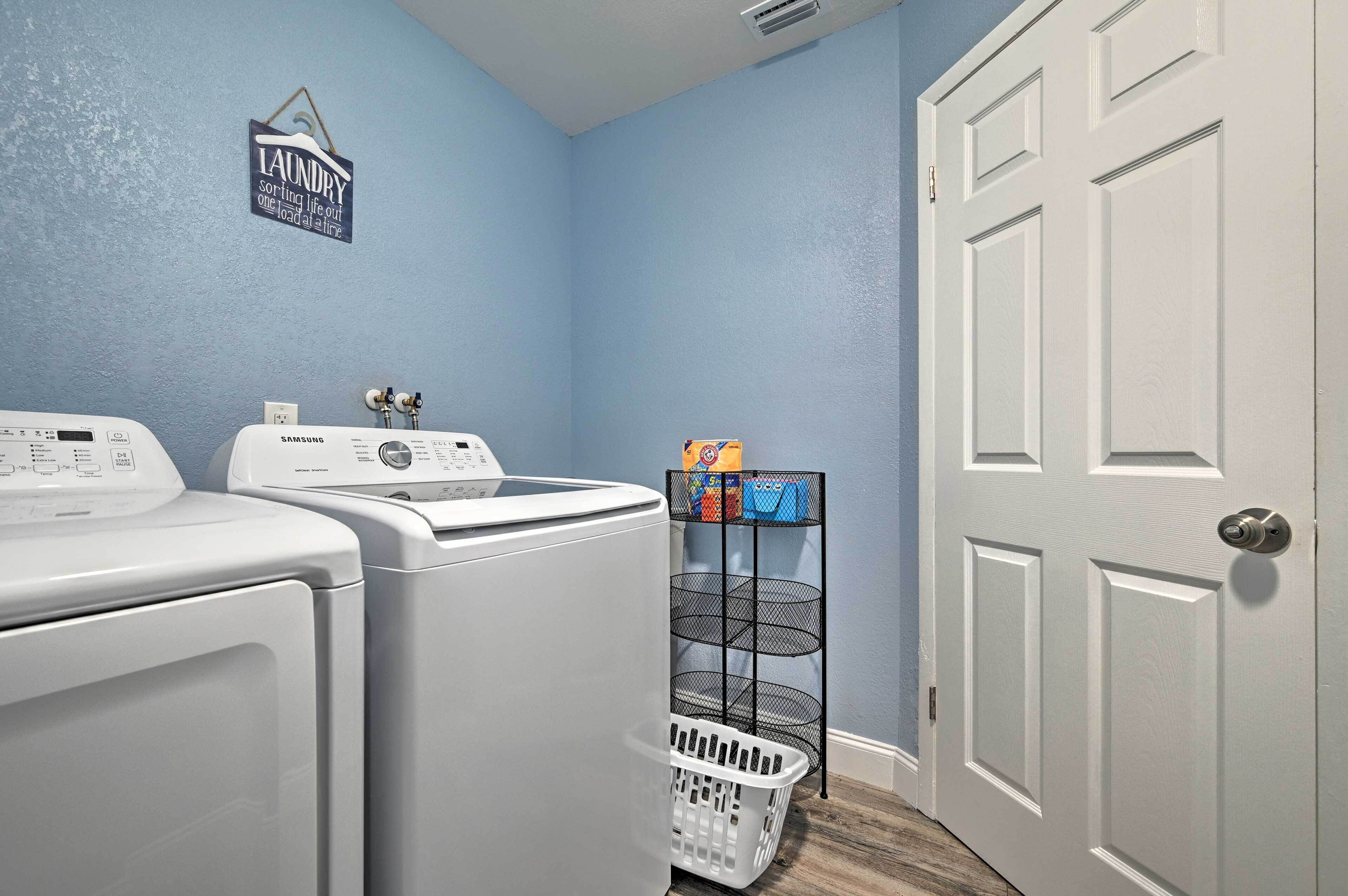 A laundry room with two front-loading washers and dryers, a metal rack with laundry baskets, and a door to the right.