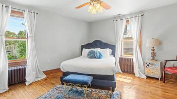 A bedroom with a bed, a ceiling fan, a window with curtains, a radiator, a rug, and a nightstand with a lamp.