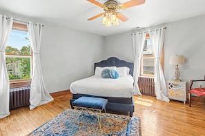 Un dormitorio con cama, ventilador de techo, ventana con cortinas, radiador, alfombra y mesita de noche con lámpara.