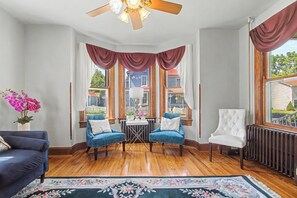 Ein Zimmer mit Holzboden, zwei blaue Sessel, ein weißer Stuhl, ein kleiner Tisch mit Blumen in einer Vase und Blick auf ein Haus durch große Fenster.