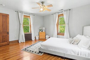 Un dormitorio con cama, mesita de noche, cómoda, ventilador de techo y dos ventanas con cortinas.