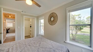 Un dormitorio con colcha estampada, ventilador de techo, ventana grande con vistas al exterior y espejo circular en la pared.