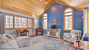 A cozy living room with a high wooden ceiling, large windows, a fireplace, and a television.