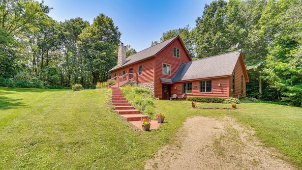 A log cabin with a stone chimney, a red exterior, and a gravel driveway leading to a wooden deck.