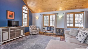A cozy living room with a wooden ceiling, a large window, a television, a sofa, and a patterned armchair.