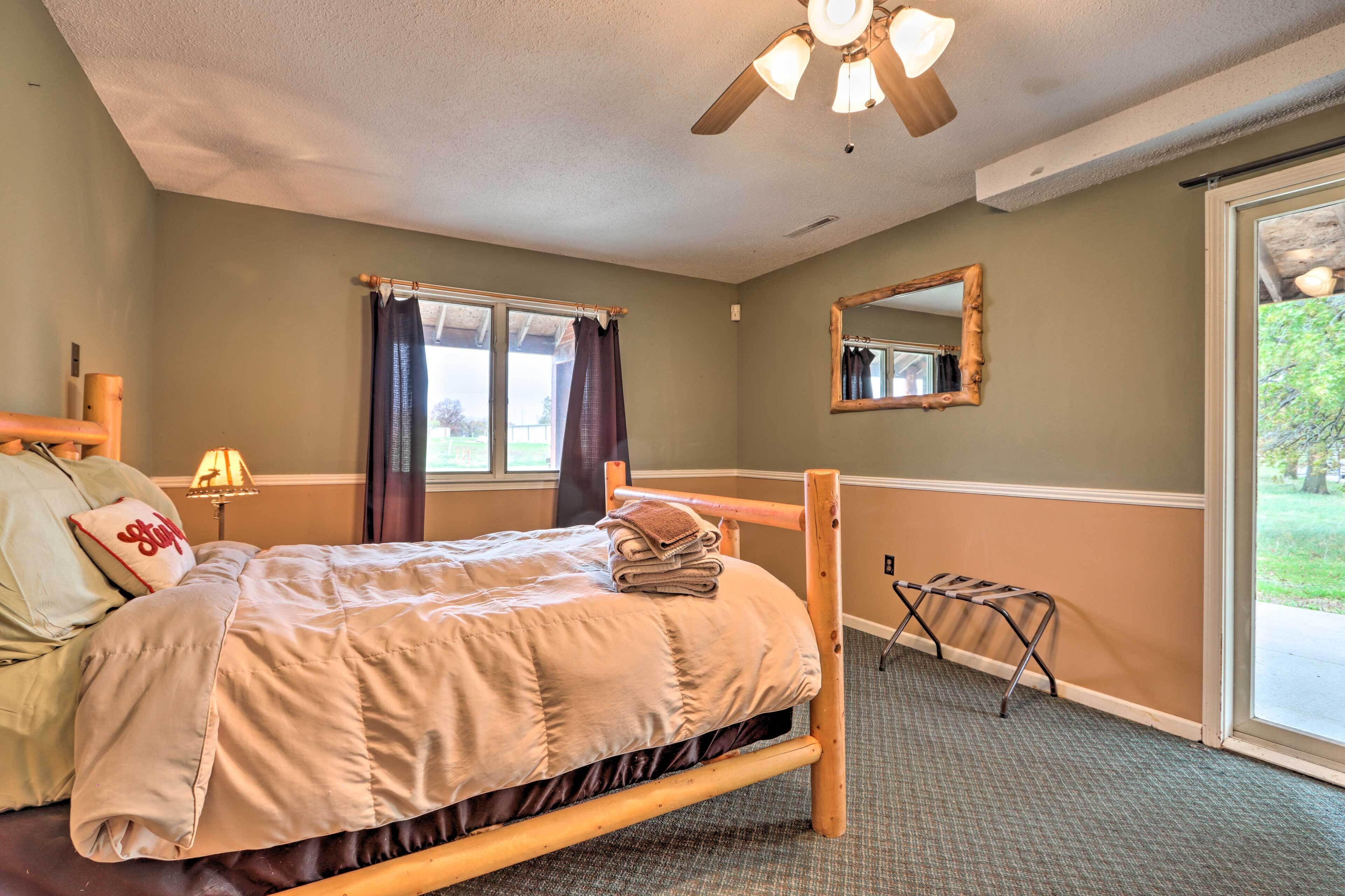 Une chambre avec un lit en bois, un ventilateur de plafond, une fenêtre avec des rideaux, un miroir et une petite table avec une lampe.