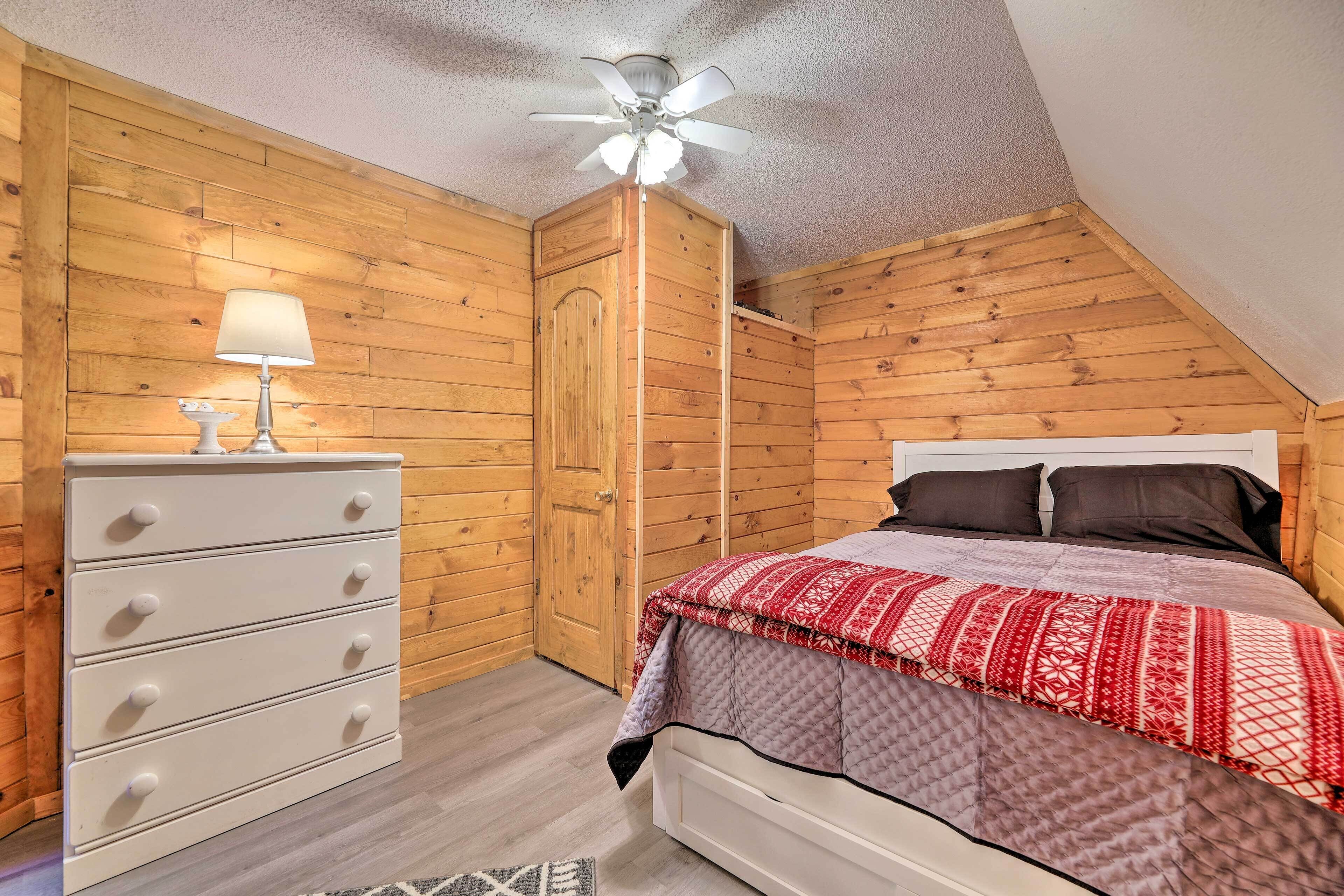 A bedroom with a wooden ceiling, a bed with a red and white patterned bedspread, a white dresser with drawers, and a ceiling fan.