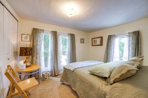 Un dormitorio con cama, una silla, una lámpara, una mesita y una ventana con cortinas.