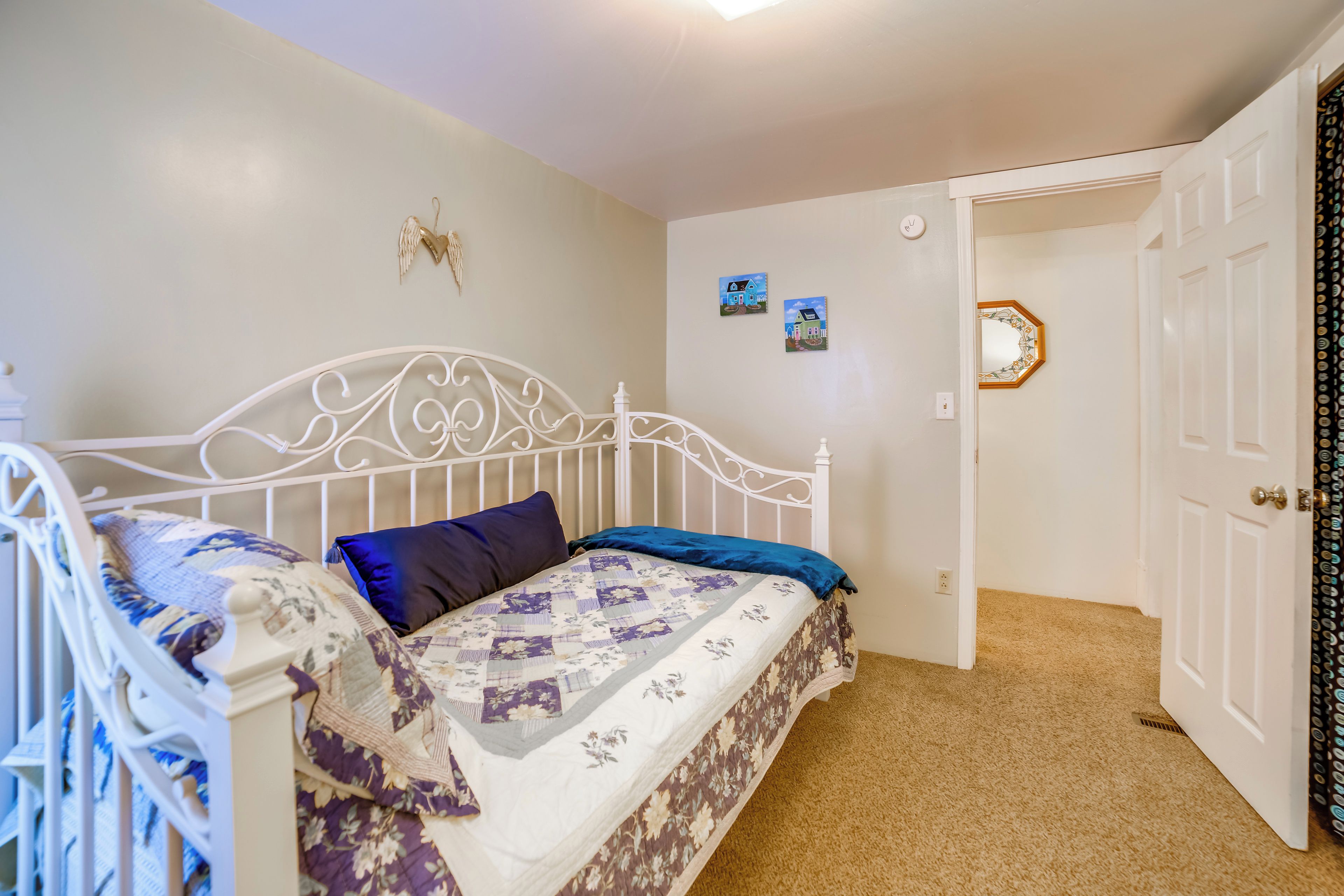 Une chambre à coucher avec un lit en métal blanc, une couvre-lit à motifs floraux et un oreiller bleu. On y trouve une petite décoration murale et une porte avec un miroir.