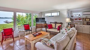 A spacious living room with a large window overlooking a body of water, a television, and a dining area with a tray of food. - Waterfront Vacation Rental on Lake Mirror! (Winter Haven)