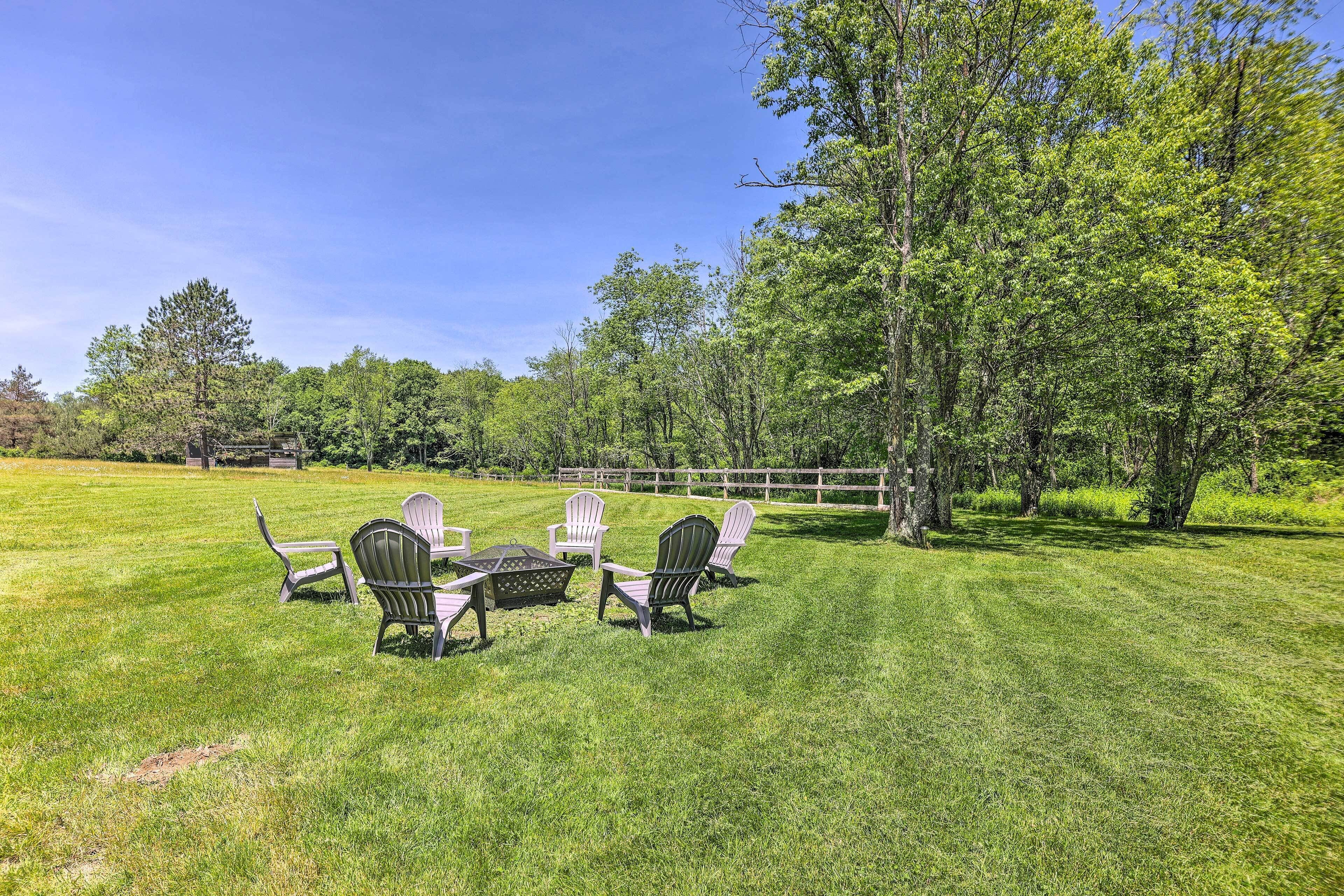 A grassy outdoor area with Adirondack chairs and a table, surrounded by trees and a wooden fence.