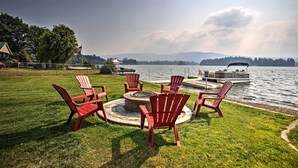A lakeside deck with red Adirondack chairs and a fire pit, overlooking a calm lake with a boat docked nearby.