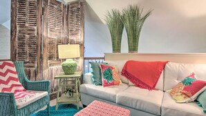 A cozy living room with a white sofa, teal armchairs, and a patterned ottoman. A wooden door with shutters and a small table with a lamp are also visible.