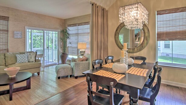 A spacious living room with a dining area, large windows, a chandelier, and a mirror.
