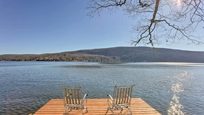 Une jetée en bois avec deux chaises en métal, donnant sur un lac paisible, le tout entouré de collines et sous un ciel dégagé.