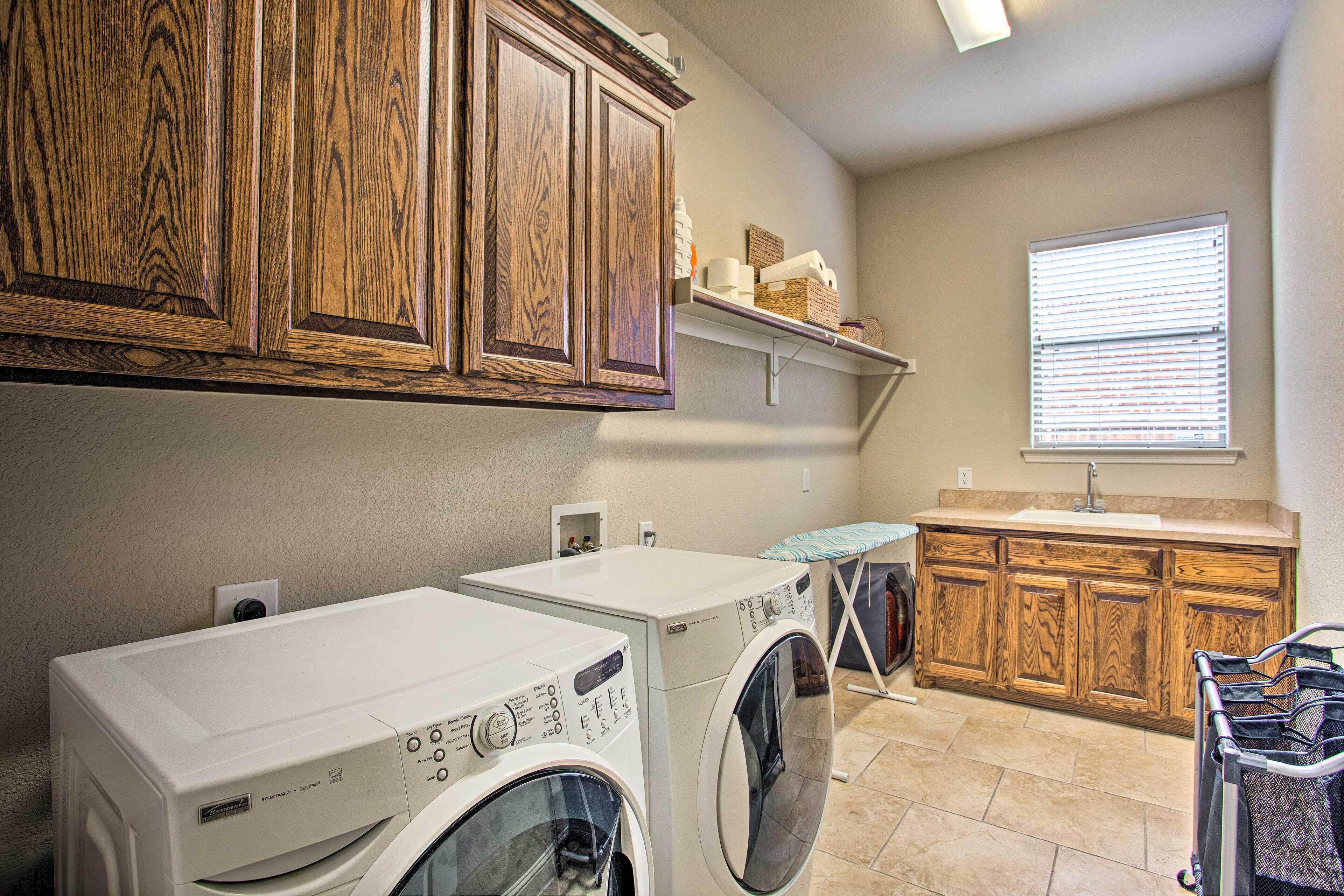 A laundry room with two front-loading washers, wooden cabinets, a countertop with a sink, and a window with blinds.