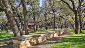 A stone bench in a park with trees and a small building in the background. - Blanco Riverfront Historic Home, 3 Mi to Downtown! (Blanco)