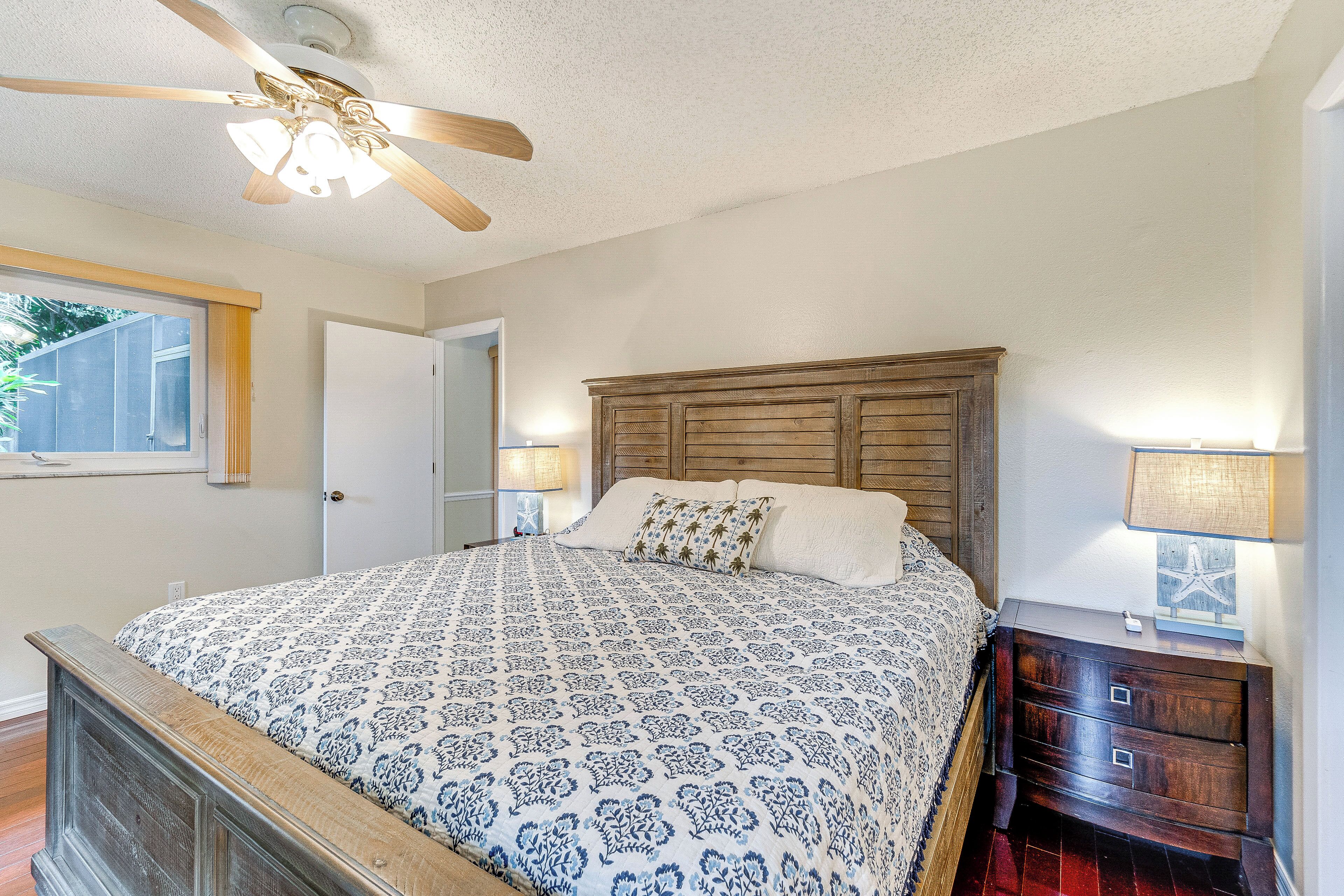 A bedroom with a wooden headboard, a patterned bedspread, a ceiling fan, and a nightstand with a lamp.