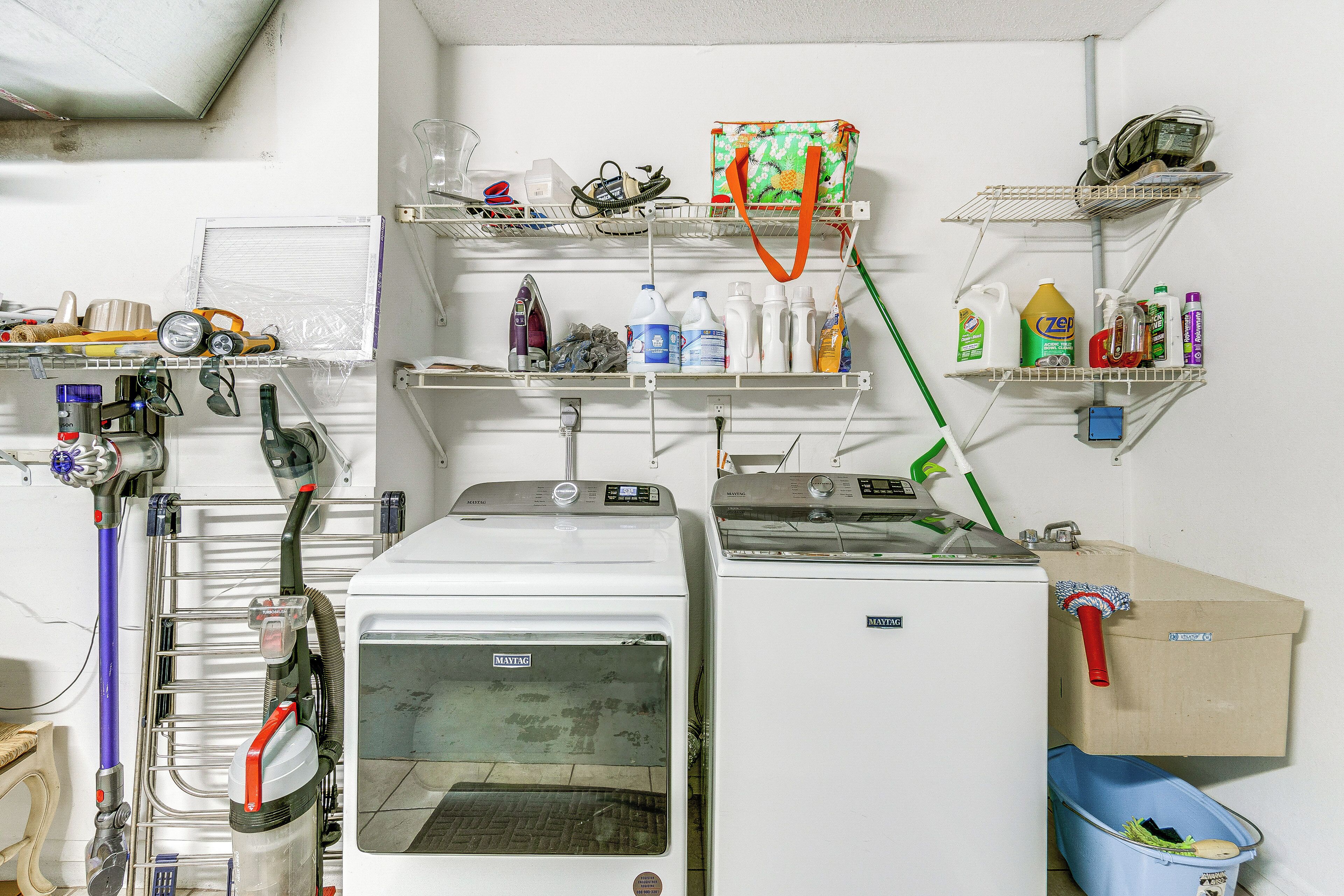 A laundry room with multiple washing machines, shelves with cleaning supplies, and a blue trash bin.