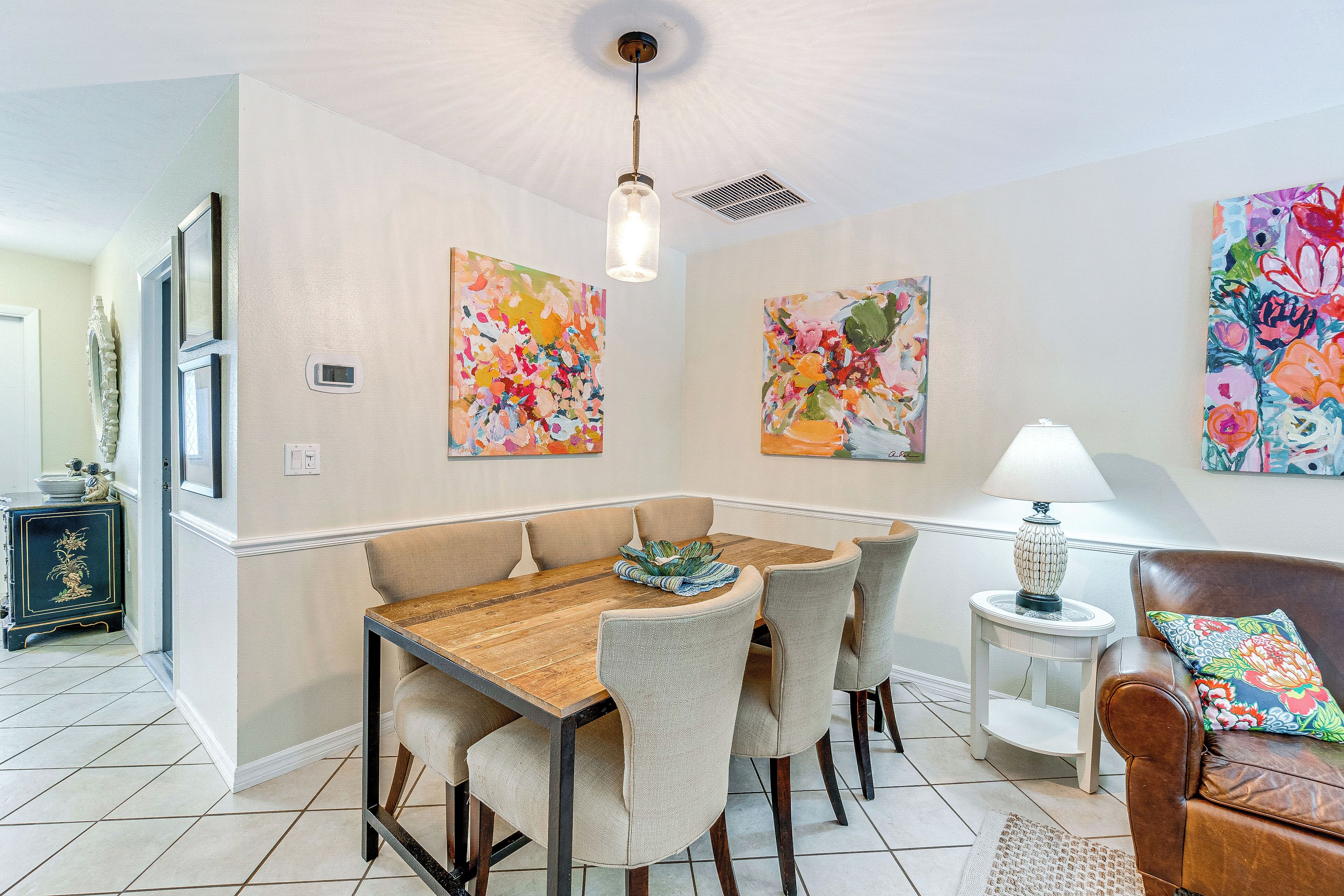 A dining area with a wooden table and chairs, a lamp, and colorful artwork on the walls.