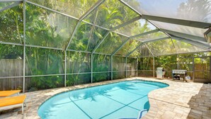 A covered swimming pool with a view of tropical foliage, a barbecue area, and a wooden fence.