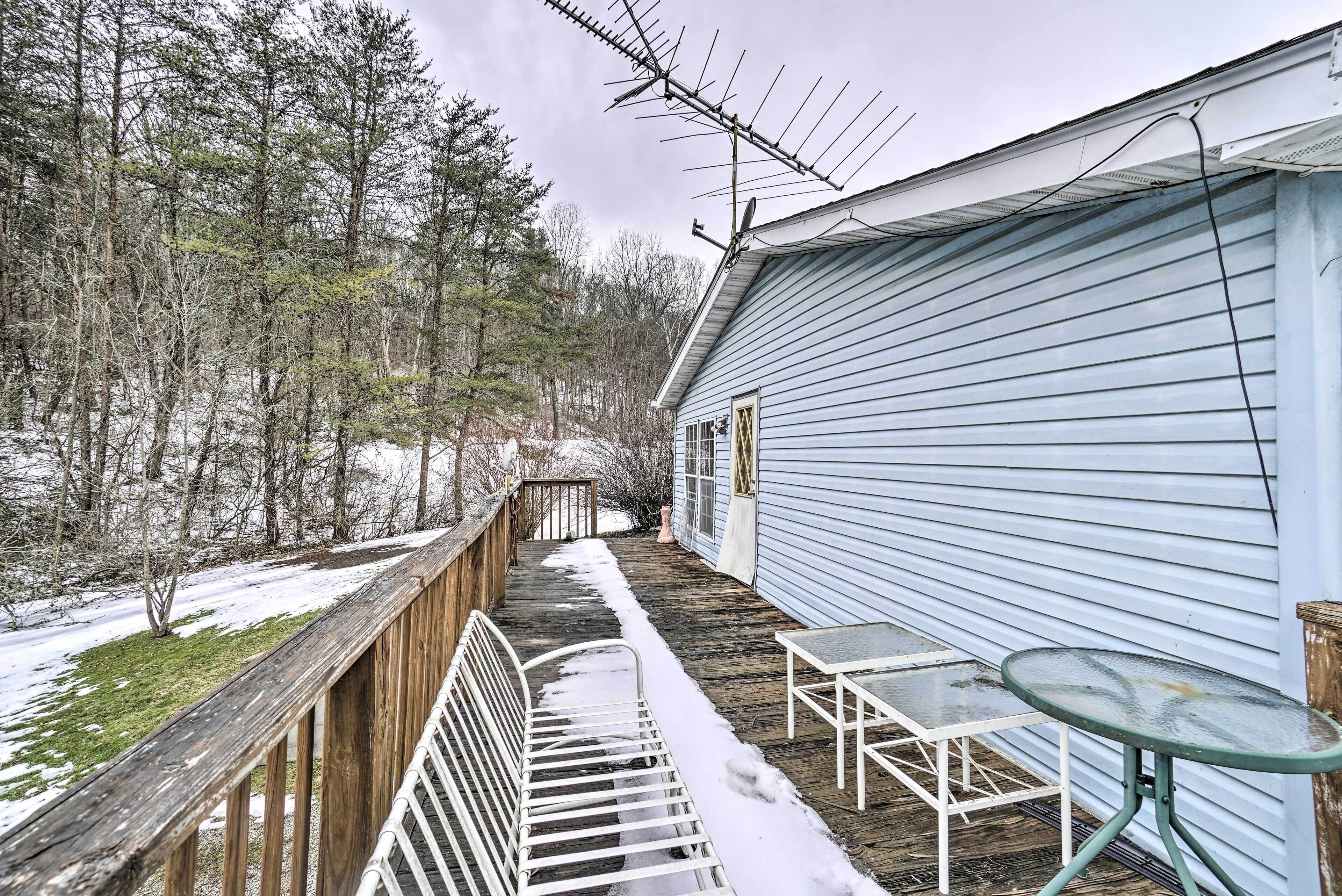 A covered outdoor deck with a metal bench and a table, surrounded by snow-covered ground and trees.