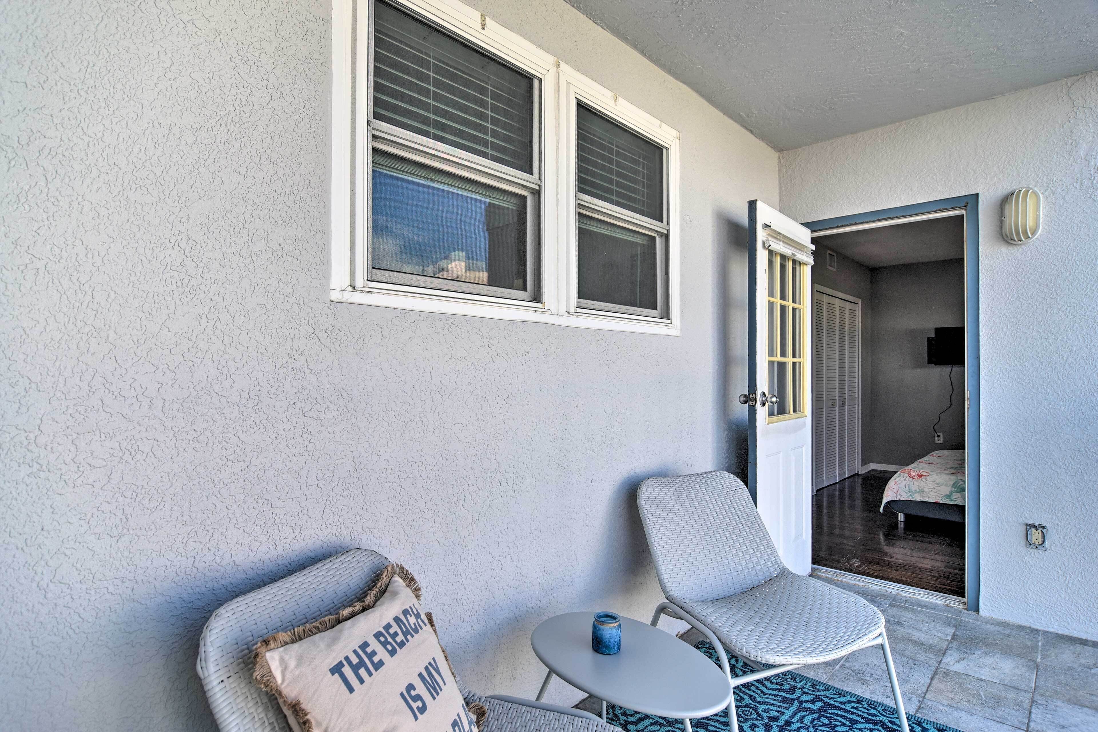 A room with a white textured wall, two windows with blinds, a wicker chair, a small round table with a blue object, and a door leading to another room.