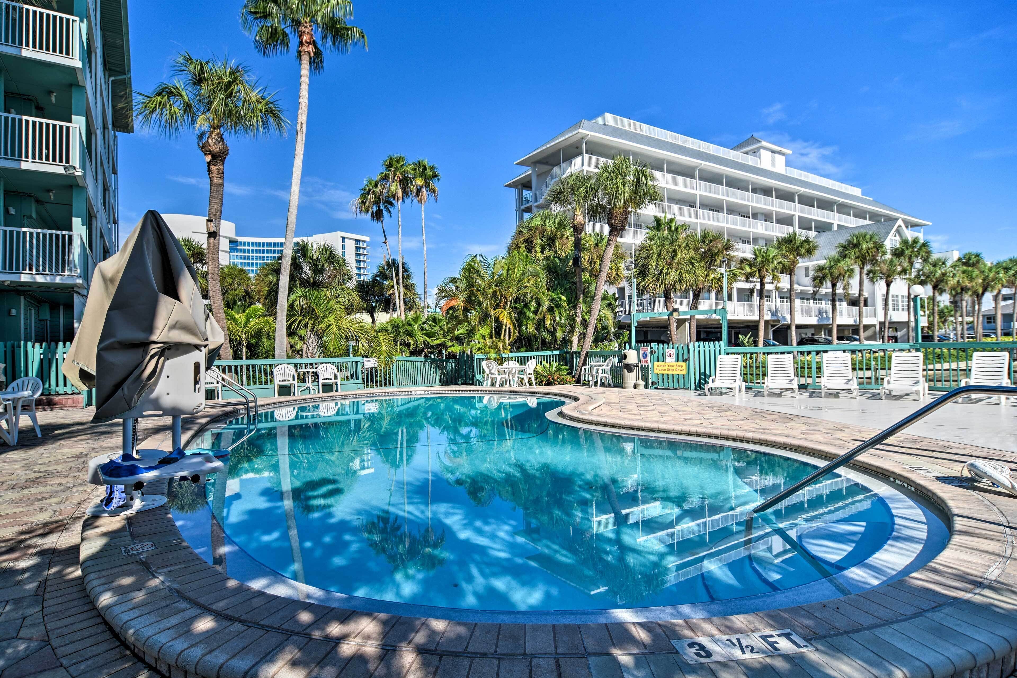 A swimming pool with a clear blue water, surrounded by palm trees and a building with multiple balconies in the background.