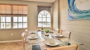 Un coin repas avec une table en verre, des chaises blanches et une grande fenêtre équipée de stores.
