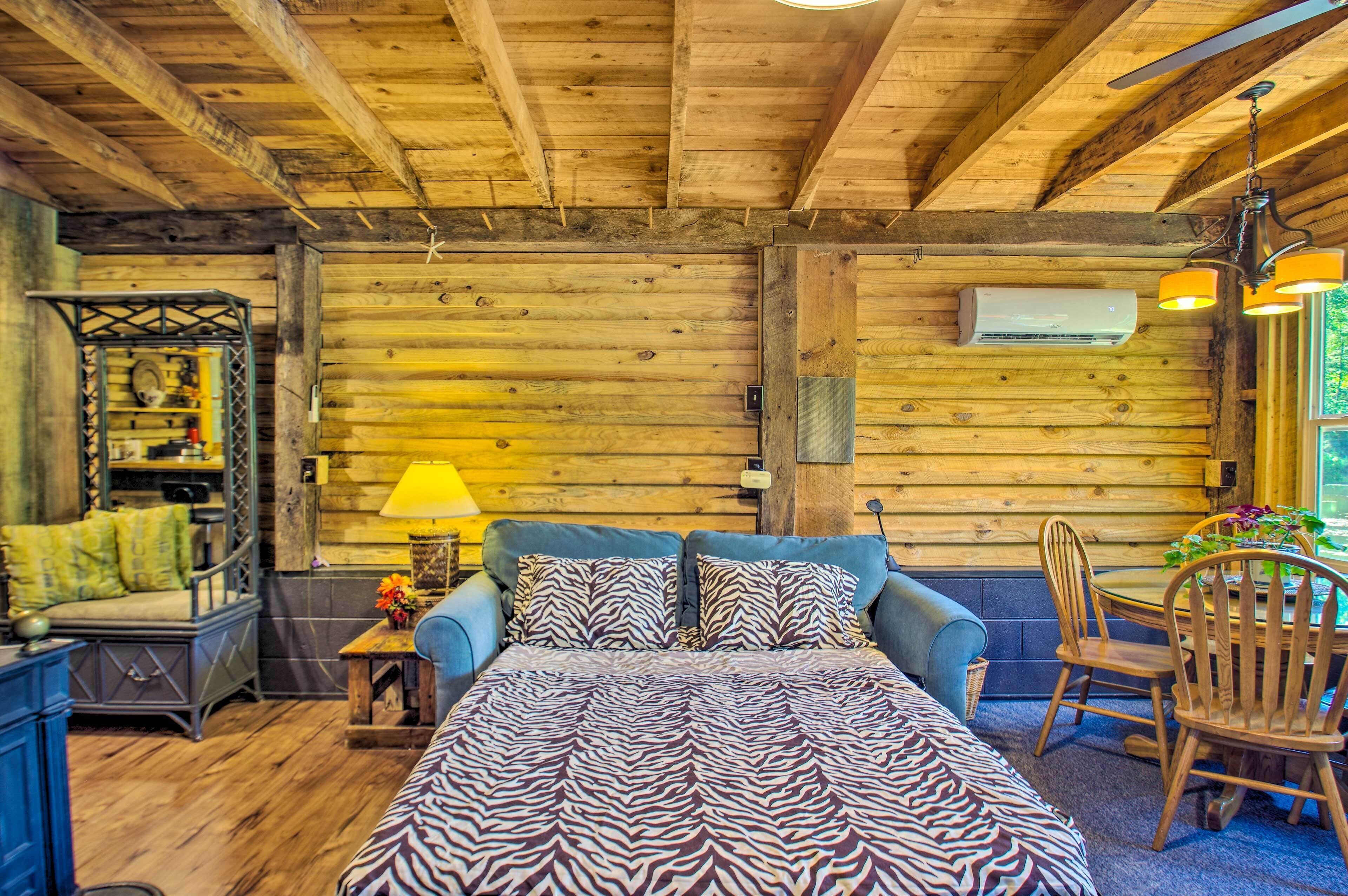 A log cabin interior with a bed, a table with chairs, a lamp, and a window with a view.