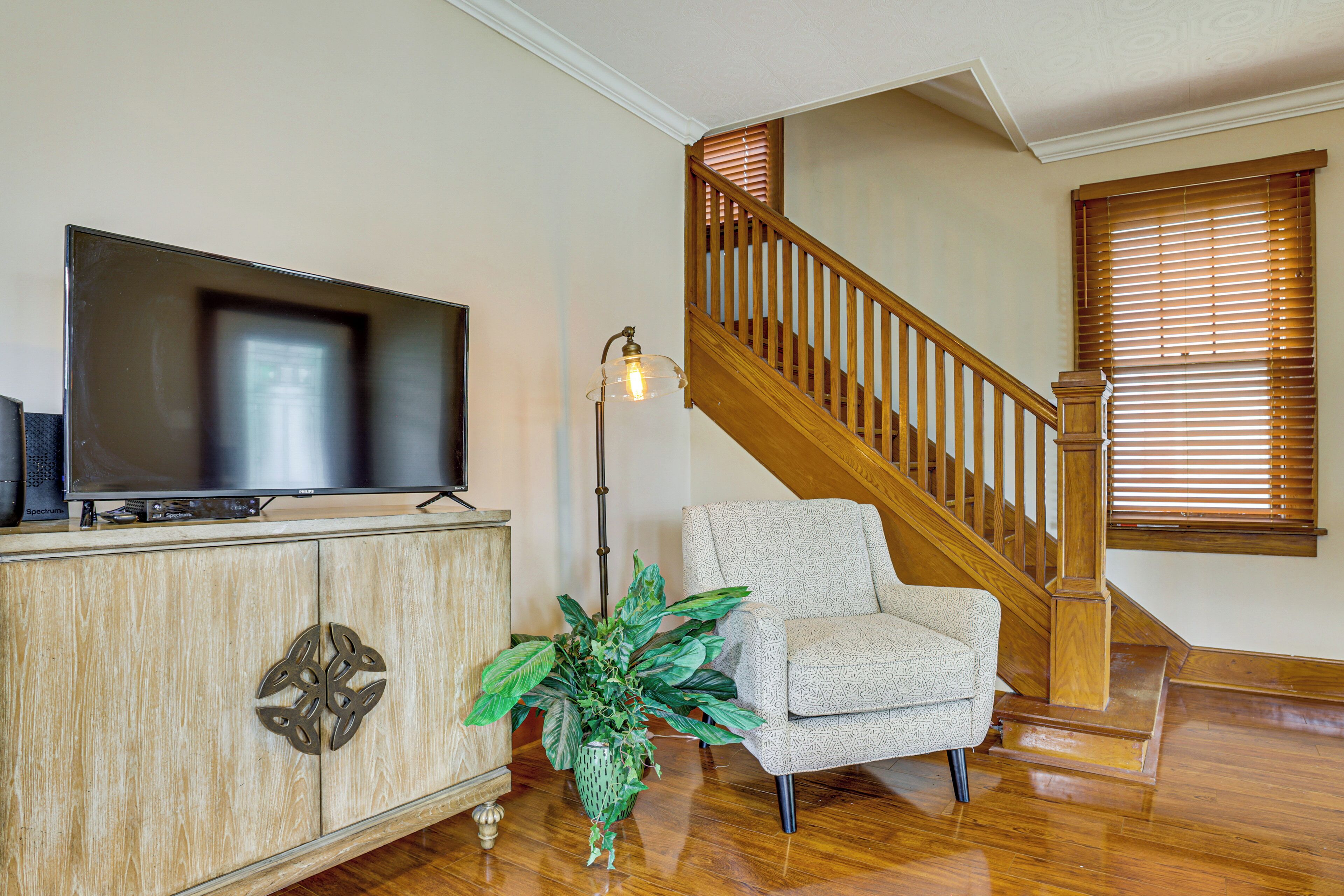 A living room with a television on a wooden cabinet, a patterned armchair, a potted plant, and a wooden staircase.