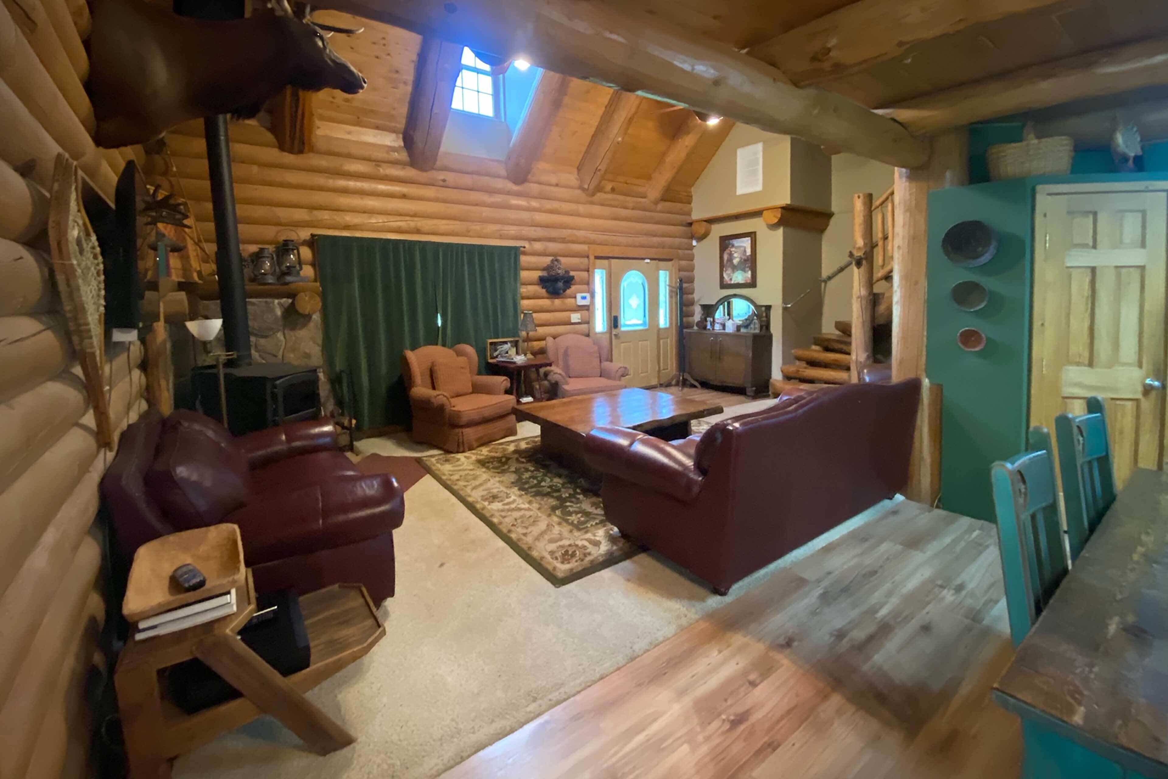 A log cabin interior with a fireplace, leather sofas, a wooden dining table, and a mounted deer head.