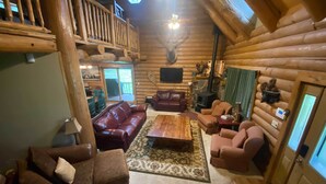 A log cabin interior with a wooden ceiling, a mounted deer head, a fireplace, and a cozy seating area with leather sofas and a coffee table. - Cozy Alto Cabin on 2 Acres: Near Fishing & Skiing! (Alto)