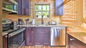 A compact kitchen with dark wood cabinets, stainless steel appliances, and a window with a view of greenery. - Mountain Dew Cabin w/ Hot Tub & Scenic Views! (Sevierville)