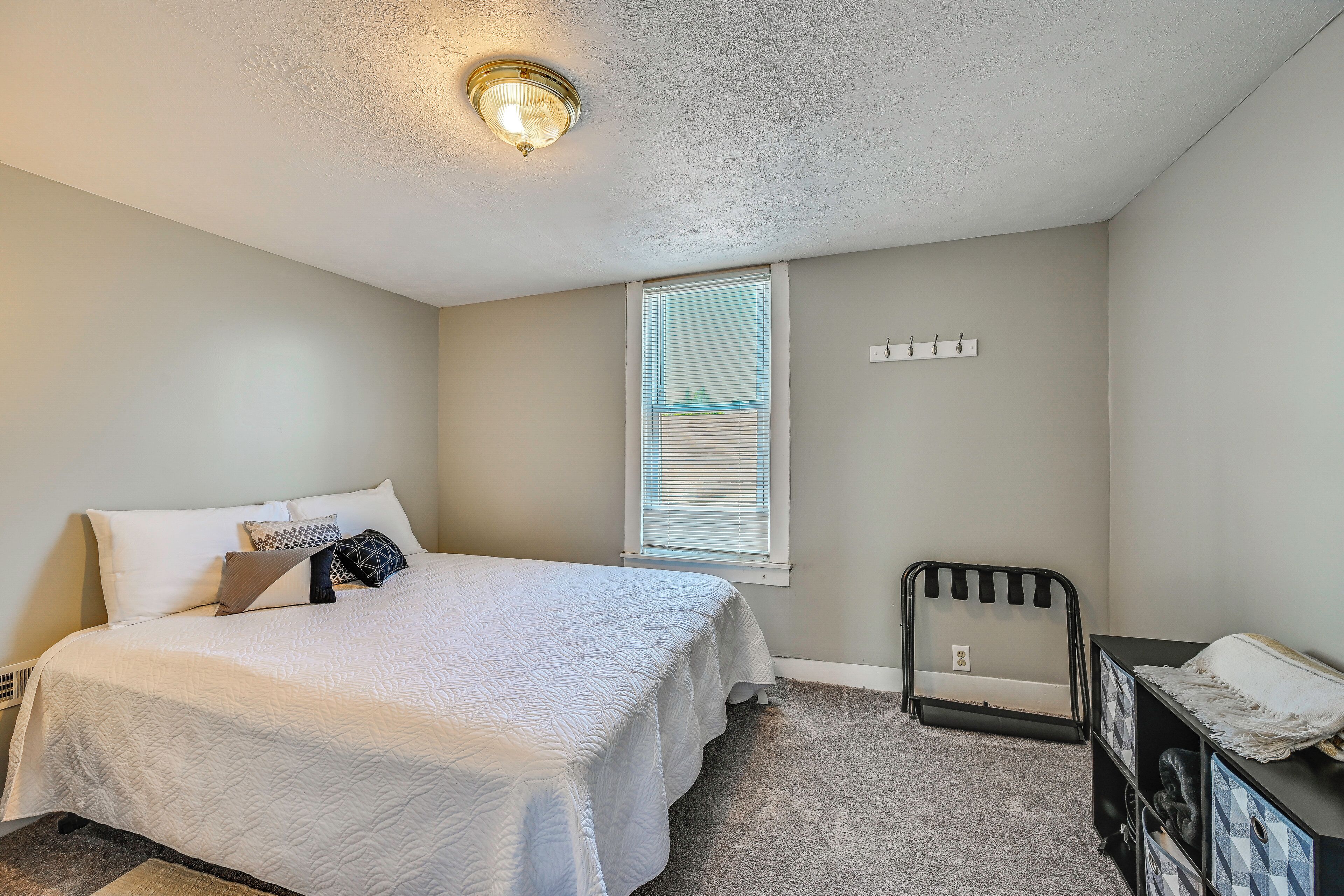 A bedroom with a bed, a window with blinds, a wall-mounted light fixture, and a black metal rack.