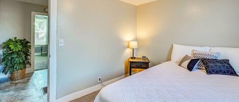 A bedroom with a bed, a nightstand, a lamp, a potted plant, and a door leading to another room.
