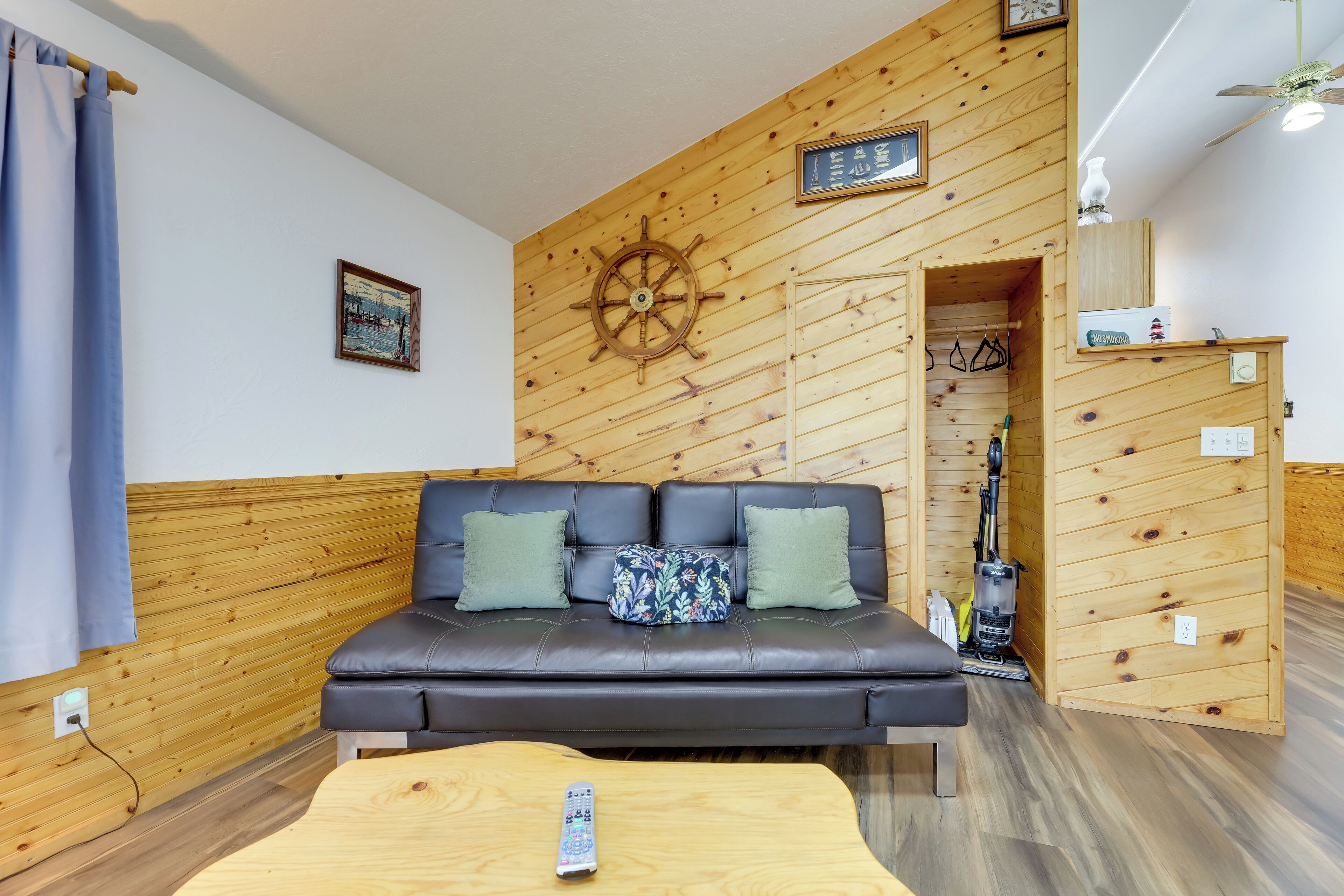 Een kamer met een houten wand, een zwarte leren bank met groene kussens, een houten salontafel en een plafondventilator.
