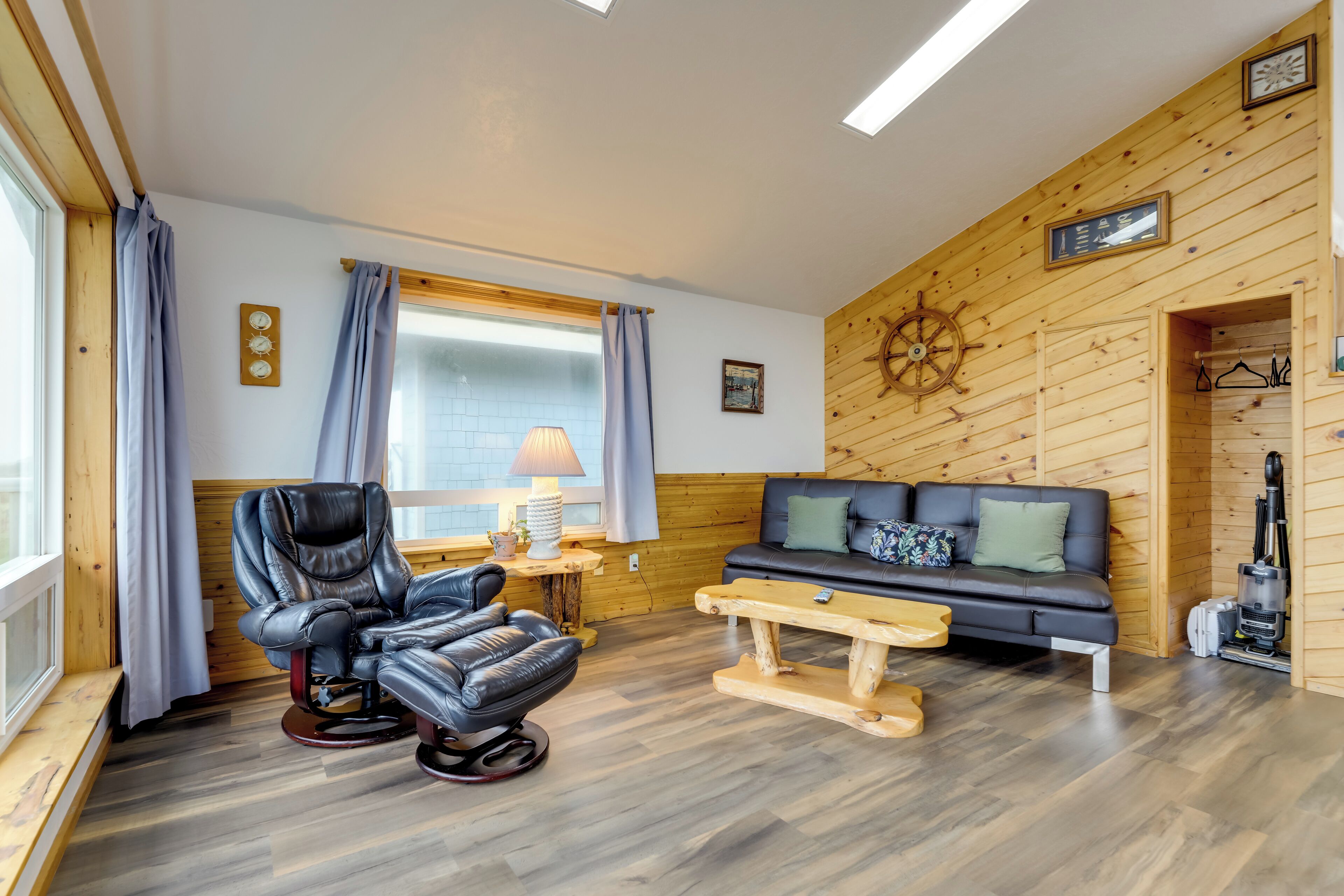 A cozy room with a leather recliner, a sofa, a wooden wall with a ship's wheel, and a window with curtains.