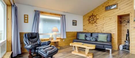 A cozy room with a leather recliner, a sofa, a wooden wall with a ship's wheel, and a window with curtains.