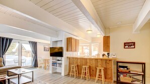 A modern living room with a sofa, coffee table, and TV, a kitchen with wooden cabinets and stools, and a bar area. - Cozy Condo Near Golf, 7 Mi to Big Sky Resort! (Big Sky)