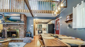 A spacious living area with a fireplace, wooden dining table, and a view of the kitchen. - Hot Tub, Fire Pit & Barrel Sauna: Poconos Abode (Long Pond)