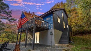 A modern house with a balcony, an American flag, and a wooden sculpture. - Hot Tub, Fire Pit & Barrel Sauna: Poconos Abode (Long Pond)
