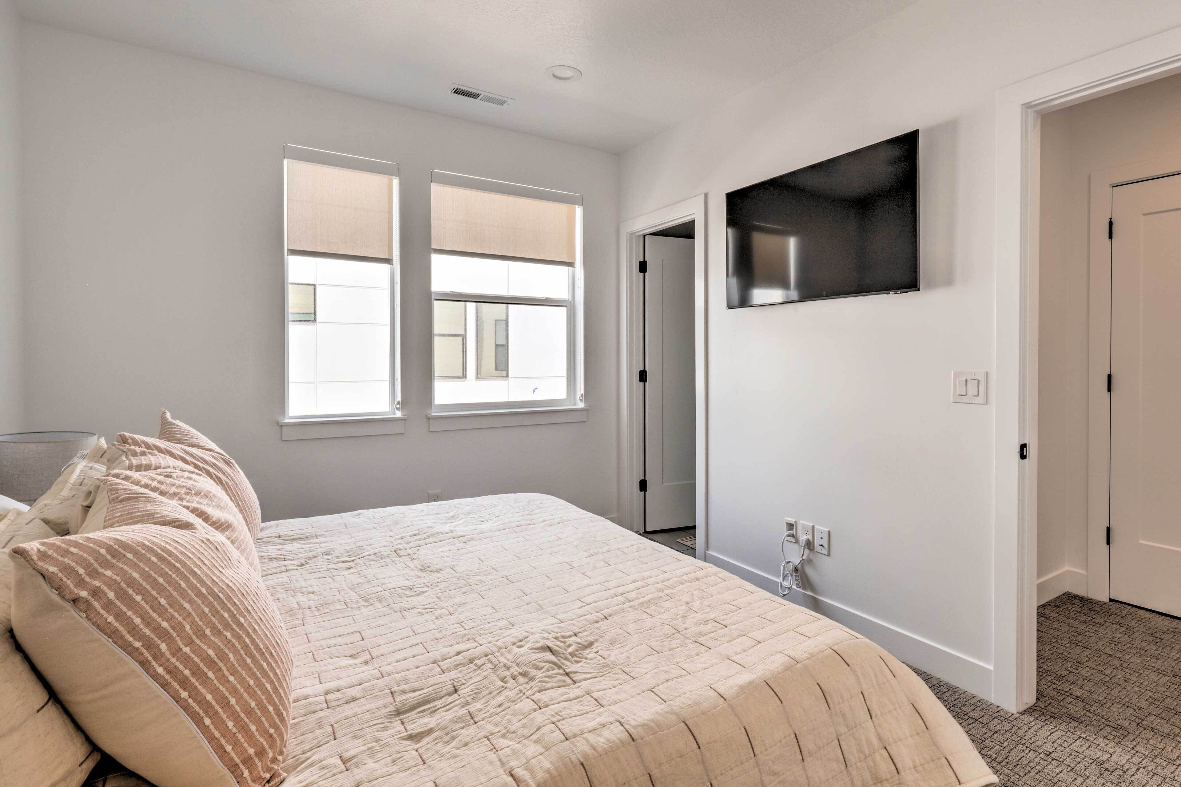 Un dormitorio con una cama, dos ventanas, un televisor montado en la pared y una puerta que da a otra habitación.