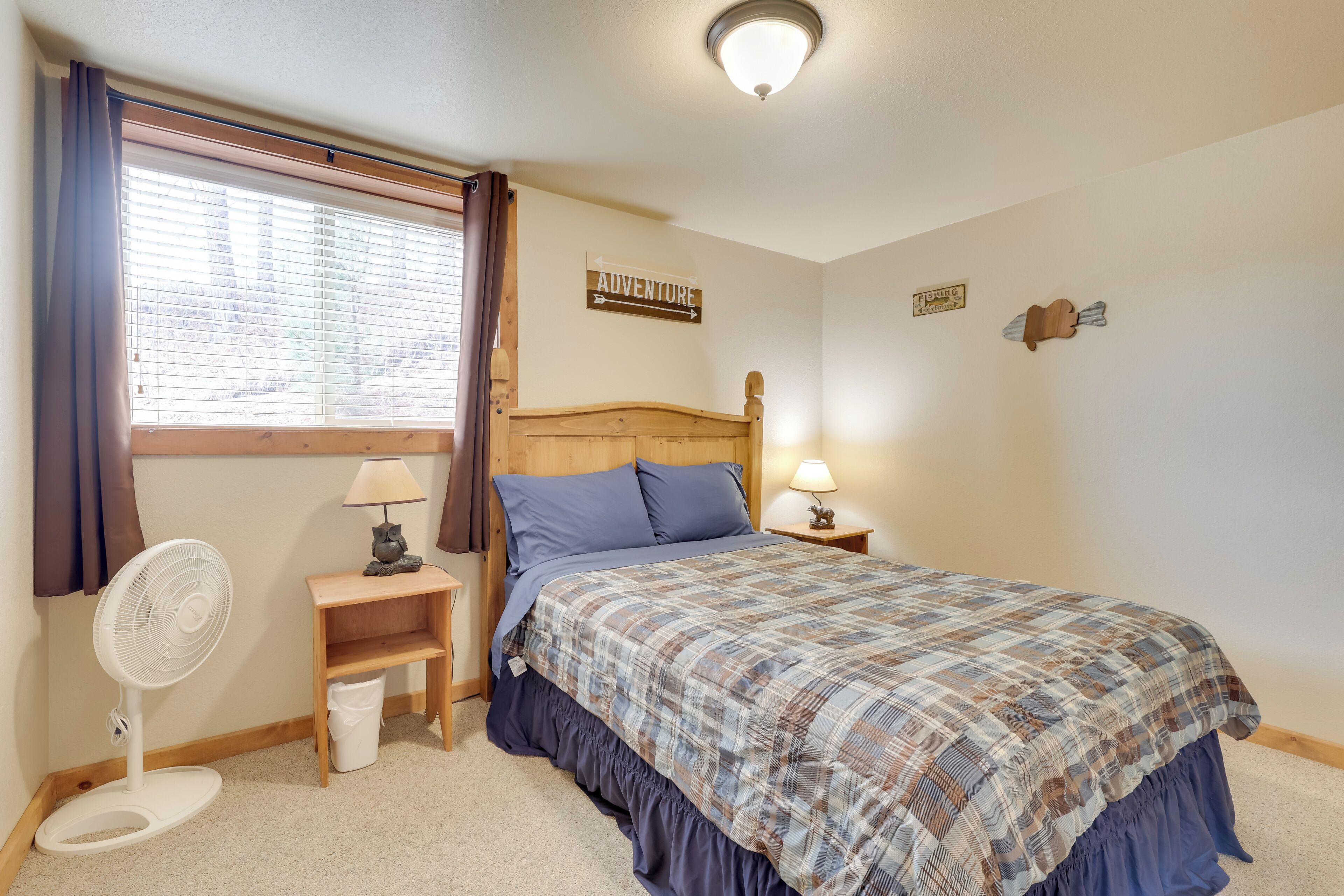 A bedroom with a bed, a nightstand, a fan, a window with curtains, and a wall sign that says "ADVENTURE".