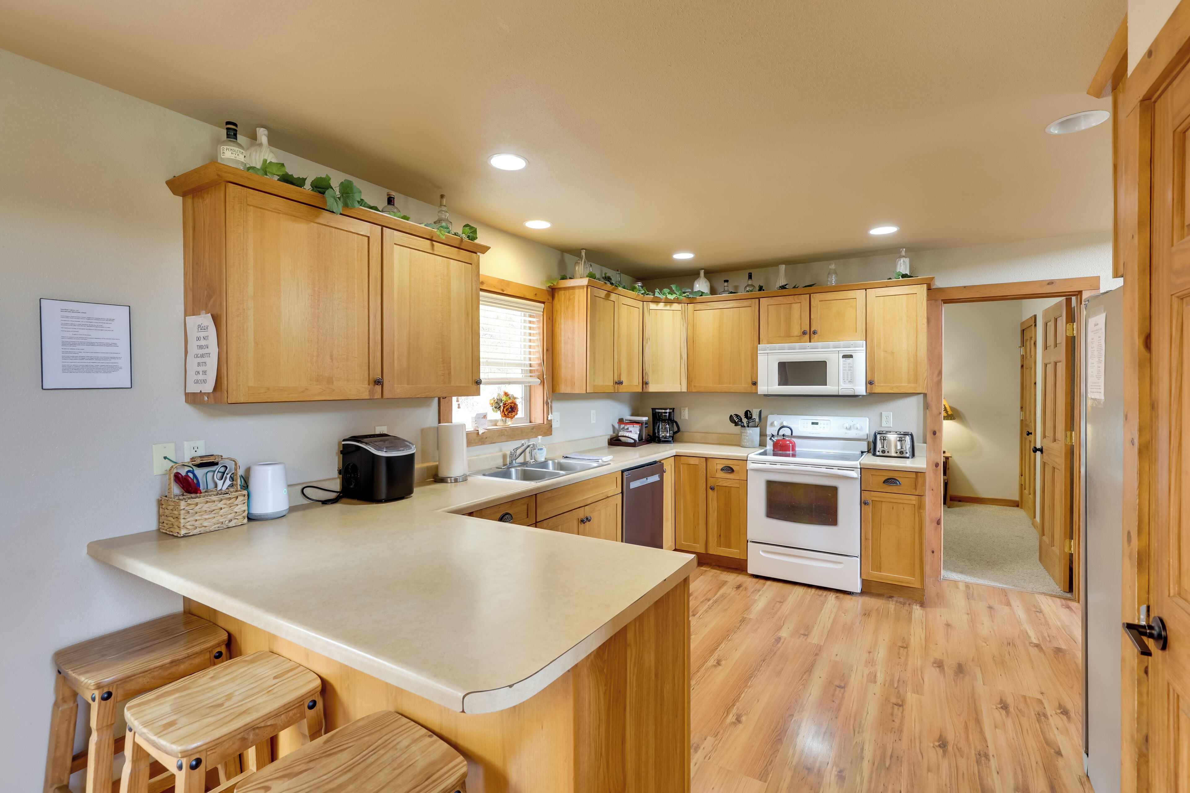 A kitchen with wooden cabinets, a microwave, a stove, and a sink. There are stools at the counter and a basket with utensils.
